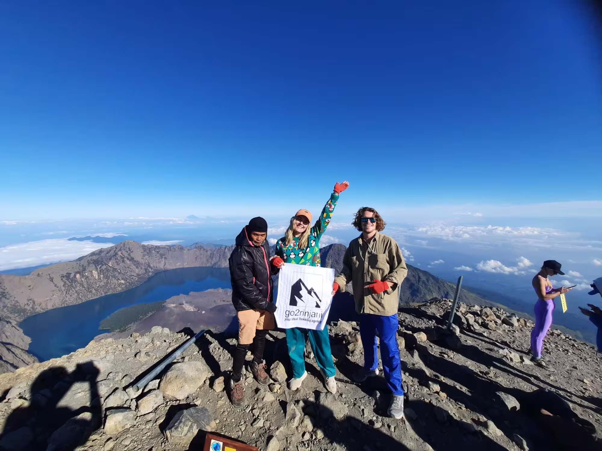A group of happy trekkers celebrating at the peak during a Mount Rinjani summit climb, holding a go2rinjani flag with the vast crater lake below.